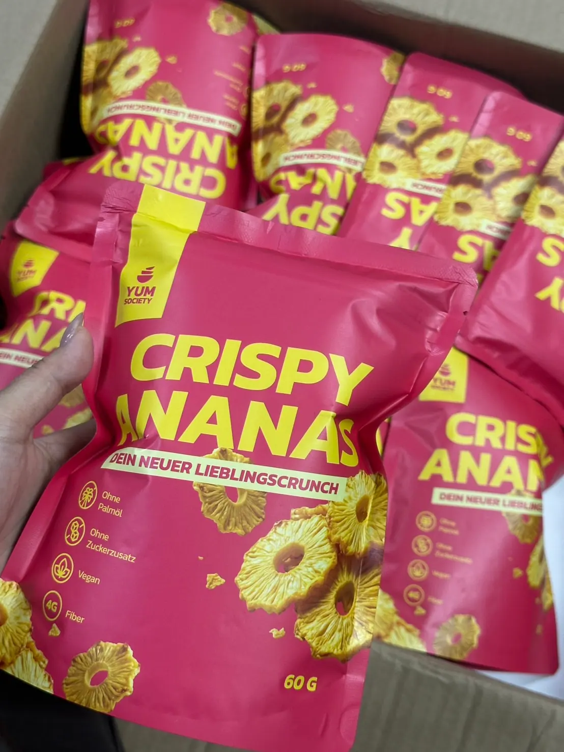 Eine Hand hält einen Crispy-Ananas-Beutel über einem Karton voller Beutel